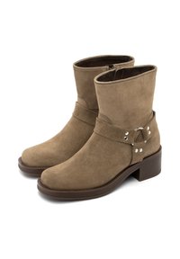 Botas de ante marrón hasta el tobillo con punta redondeada, un tacón plano y un detalle de correa con herrajes de plata. Textura suave y diseño simple.