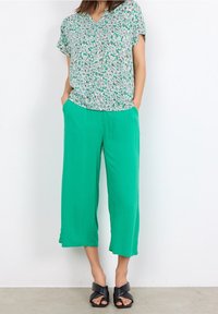 Bluse med blomsterprint, kortærmet i hvid og grøn, parret med grønne, vide bukser. Sorte sandaler med åbne tæer fuldender looket.