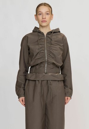 Femme aux cheveux blonds portant un sweat à capuche marron froncé zippé et un pantalon ample assorti sur un fond clair uni.
