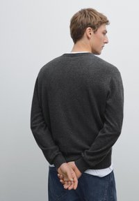 Maglione grigio scuro con scollatura rotonda e polsini a costine, indossato sopra una t-shirt bianca, con una vestibilità comoda e una texture liscia.