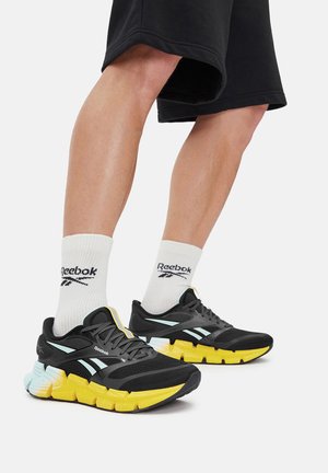 Czarno-żółte sportowe buty z siateczkową tkaniną, grubą podeszwą i jasnoniebieskimi akcentami; w połączeniu z białymi skarpetkami z logo.