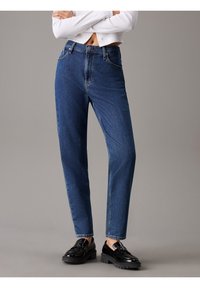 Person som bär högmidjade blå jeans och svarta loafers, med armarna i kors och en kortärmad vit tröja mot en enfärgad bakgrund.