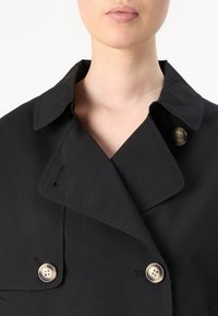 Cappotto trench nero con ampio colletto, caratterizzato da grandi bottoni metallici e una texture liscia. Design doppiopetto con dettagli di cucitura.