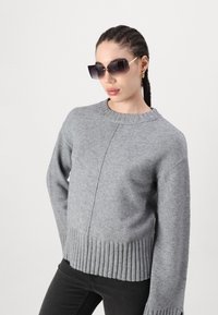 Grijze gebreide trui met geribbelde onderrand en manchetten, met een ronde hals en een gladde textuur. Model draagt een oversized, donkere zonnebril.