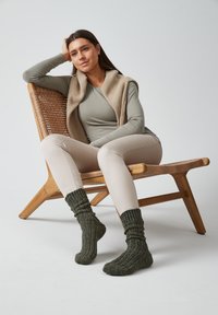 Gilet in maglia beige, maglia a maniche lunghe verde oliva, pantaloni beige chiaro e calze verdi a coste. Seduto su una sedia di legno con sedile intrecciato.