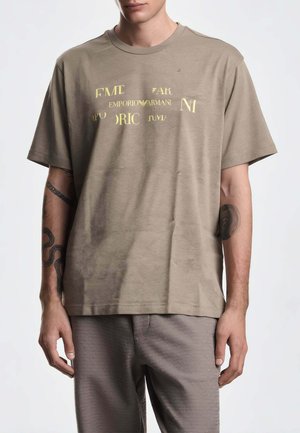 Uomo che indossa una t-shirt beige Emporio Armani con scritta dorata, mostrando tatuaggi su entrambi gli avambracci, in piedi davanti a uno sfondo semplice.
