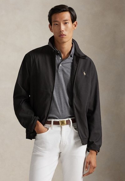 Man draagt een zwarte jas met ritssluiting over een grijs poloshirt en witte broek, staat met één hand in de zak tegen een effen achtergrond.