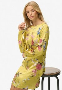 Geel satijnen jurk met lange mouwen, bloemenpatronen in roze en blauw, ronde halslijn en een losse fit, gedragen terwijl ze op een kruk zit.