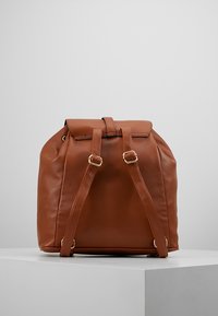 Brauner Leder-Rucksack mit strukturiertem Material, umklappbarem Deckel und verstellbaren Trägern. Verfügt über Hardware in Goldtönen und einen abgerundeten Boden.