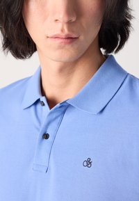 Tricou polo de culoare albastru deschis, realizat dintr-un material texturat, având un guler clasic, două nasturi negri și un mic logo negru pe piept.