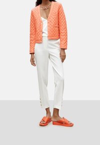 Vadderad koralljacka med knäppning, vita byxor med knapputstickare, och orange sandaler med tjock sula och korsremdesign.