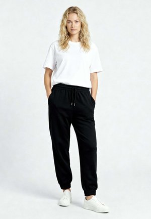 Fiatal nő göndör szőke hajjal, kezét zsebében tartva, fehér pólót, fekete joggingnadrágot és fehér tornacipőt visel egyszínű háttér előtt.