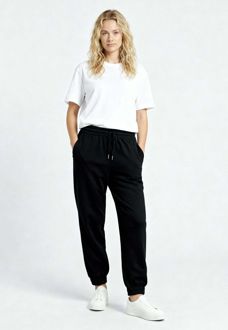 Jeune femme aux cheveux blonds bouclés, debout les mains dans les poches, portant un t-shirt blanc, un pantalon de jogging noir et des baskets blanches sur un fond uni.