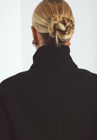 Sort kravejakke med en blød tekstur. Frisuren har en snoet knold. Sølv hoopsøreringe synlige. Minimalistisk baggrund.