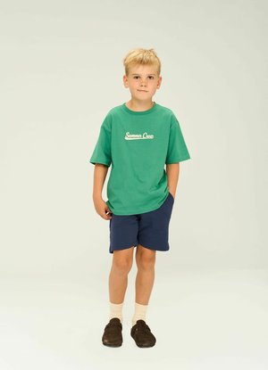 Grünes Baumwoll-T-Shirt mit dem Aufdruck "Summer Crew", marineblaue Shorts, beige Socken und dunkelbraune Schuhe. Freizeitoutfit für Kinder.