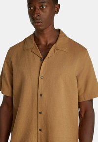 Camicetta in lino marrone a maniche corte con colletto, cinque bottoni neri e un logo sottile sul lato sinistro. Tessuto liscio, vestibilità comoda.