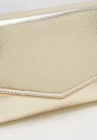 Zblízka zlatá texturovaná kabelka se zaklopem ve tvaru chevron a jemným metalickým lemem podél okraje.