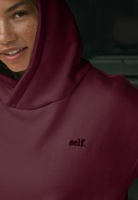 Burgundin värihuppu suurella, pehmeällä hupulla. Rinnassa brodeerattu "self." -logo. Tasainen pinta ja rento muotoilu.