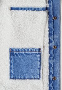 Tejido de forro polar blanco texturizado con un bolsillo a parche de denim azul y ribete, que presenta dos botones a presión de metal en el borde derecho.