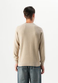Uomo di spalle che indossa un maglione beige a trama e jeans blu larghi con grandi tasche posteriori, davanti a uno sfondo bianco.