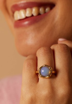Smiling person with tan skin showing a gold ring with a round, iridescent blue-purple stone on their finger against a neutral background.