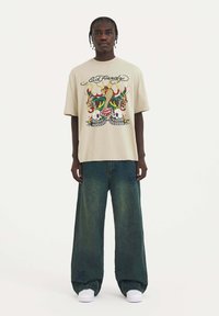 Camiseta gráfica beige con ilustraciones coloridas, combinada con unos vaqueros oscuros de corte holgado. Modelo llevando zapatillas blancas.