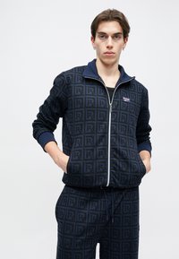 Jūras spēku zilas krāsas zip-funkcijas sporta tērps ar visā virsmā esošu logo rakstu, ar ribotām aprocēm un apkakli, divām sānu kabatām un pieguļošu griezumu.