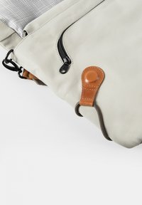 Borsa impermeabile beige con chiusura a zip, hardware nero e dettaglio in pelle marrone con bottone circolare. Texture liscia, design strutturato.