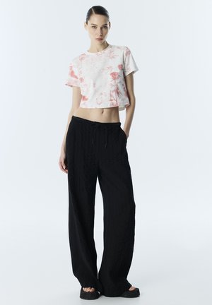 Jonge vrouw met een witte cropped top met rode bloemenprint, losse zwarte broek met trekkoord en zwarte platformsandalen, staand tegen een witte achtergrond.