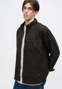 Cămașă maro cu carouri, dintr-o țesătură texturată, cu un design cu nasturi, buzunar pe piept cu patch logo, purtată peste un tricou de culoare deschisă.