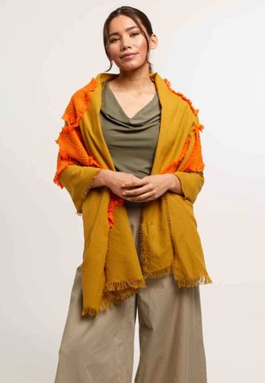 Una donna che indossa un top verde oliva, pantaloni beige a gamba larga e una sciarpa gialla senape con frange arancioni, in piedi contro uno sfondo neutro.