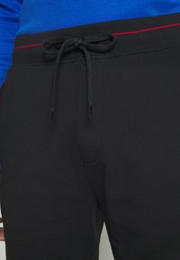 Joggers noirs en tissu doux, avec une taille à cordon rouge et des poches latérales, associés à un shirt bleu à manches longues.