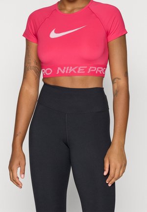 Donna che indossa un crop top Nike Pro rosa e leggings neri a vita alta, mostrando tatuaggi su entrambe le braccia su uno sfondo semplice.