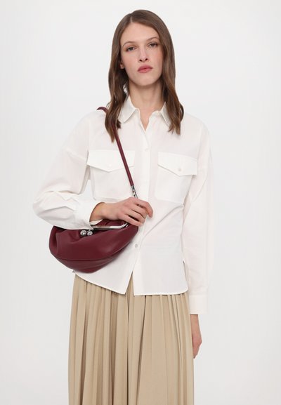 Chemise blanche à boutons avec deux poches poitrine, associée à une jupe plissée beige. Tenant un sac en cuir bordeaux avec des garnitures métalliques.