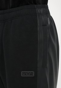 As calças de moletom pretas apresentam uma mistura de texturas de tecido, com um painel lateral em couro liso e uma pequena placa de marca no lado esquerdo da coxa.