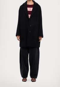 Manteau en laine noire à coupe oversize, col cranté, trois boutons et grandes poches ; porté sur un haut rayé rose et bordeaux et un pantalon noir.