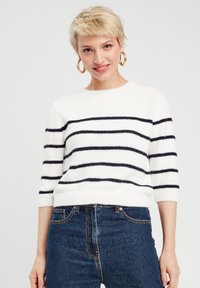 Maglione bianco a righe con strisce orizzontali blu, collo a giro, maniche a lunghezza gomito e una texture morbida e soffice abbinato a jeans in denim blu.