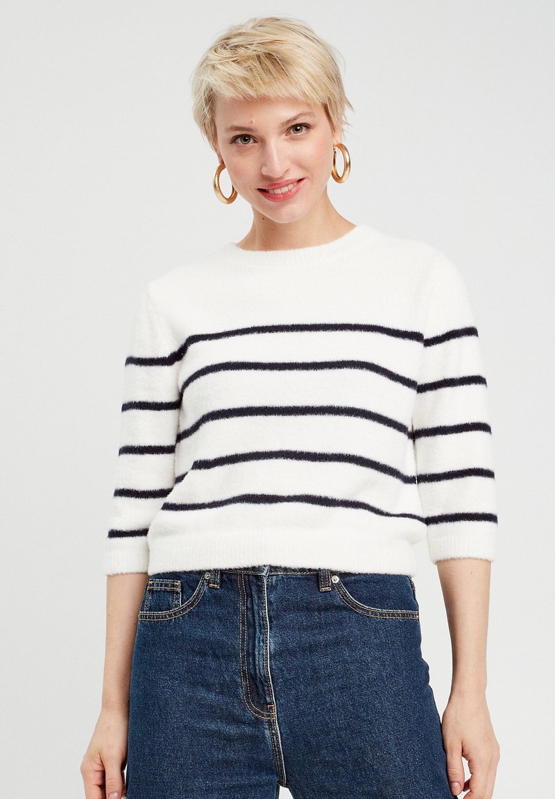 Weißer Pullover mit navyblauen horizontalen Streifen, Rundhalskragen, Ärmel bis zum Ellenbogen und weicher, flauschiger Textur, kombiniert mit blauen Jeans.