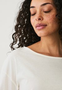 Witte, lichte top met een brede halslijn en een ontspannen pasvorm. Krullend haar en gezicht met sproeten zijn zichtbaar, met de nadruk op de gladde textuur van de stof.