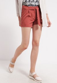 Pantaloni corti di colore ruggine in tessuto leggero con lacci in vita e tasche laterali. Abbinati a scarpe Mary Jane bianche.