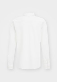 Vit långärmad skjorta med knäppta manschetter, sedd från baksidan mot en enkel ljus bakgrund.