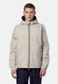 Jeune homme portant une veste à capuche beige clair fermée par une fermeture éclair, avec des poignets élastiques, debout devant un fond blanc uni.