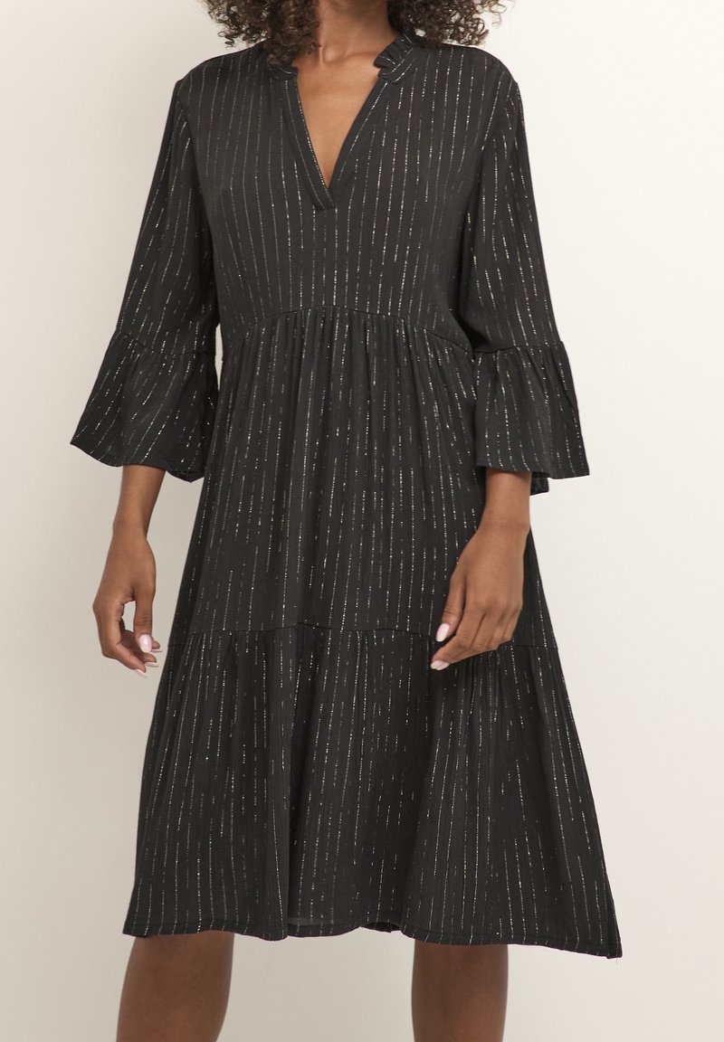 Femme portant une robe midi noire avec des rayures verticales argentées, un col en V, des manches évasées 3/4 et une jupe à volants.
