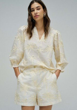 Mujer con conjunto a juego de blusa y pantalones cortos crema con patrón texturizado de palmera, manos en los bolsillos, fondo neutro.