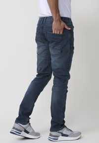 Jeans de mezclilla en lavado azul oscuro con corte ajustado, que cuentan con múltiples bolsillos y costuras en contraste. Combinados con zapatillas grises.
