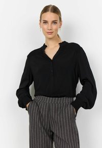 Blusa negra con cuello volante, tapeta de botones y detalle de hombros fruncidos. Mangas largas con puños fruncidos. Combinada con pantalones de rayas.