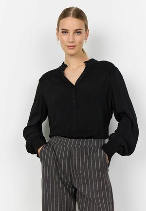 Black blouse with ruffled collar, button placket, and smocked shoulder detail. Long sleeves with gathered cuffs. Paired with pinstriped trousers.
