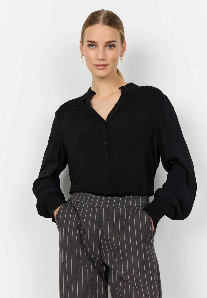 Blusa negra con cuello volante, tapeta de botones y detalle de hombros fruncidos. Mangas largas con puños fruncidos. Combinada con pantalones de rayas.