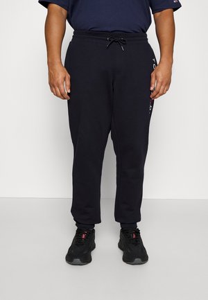 Man die zwarte joggingbroek met aantrekkoord in de taille en zwarte sneakers draagt, staand tegen een effen lichte achtergrond.