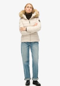 Giacca puffer color beige chiaro con cappuccio foderato in pelliccia, chiusura con zip, design trapuntato, abbinata a jeans blu e stivaletti neri.